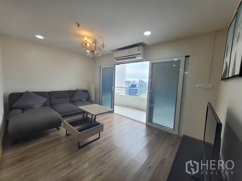 公寓 出租在Watthana, Bangkok - 明亮客厅，灰色转角沙发与水晶吊灯，连接阳台可赏通罗一带城市景观。