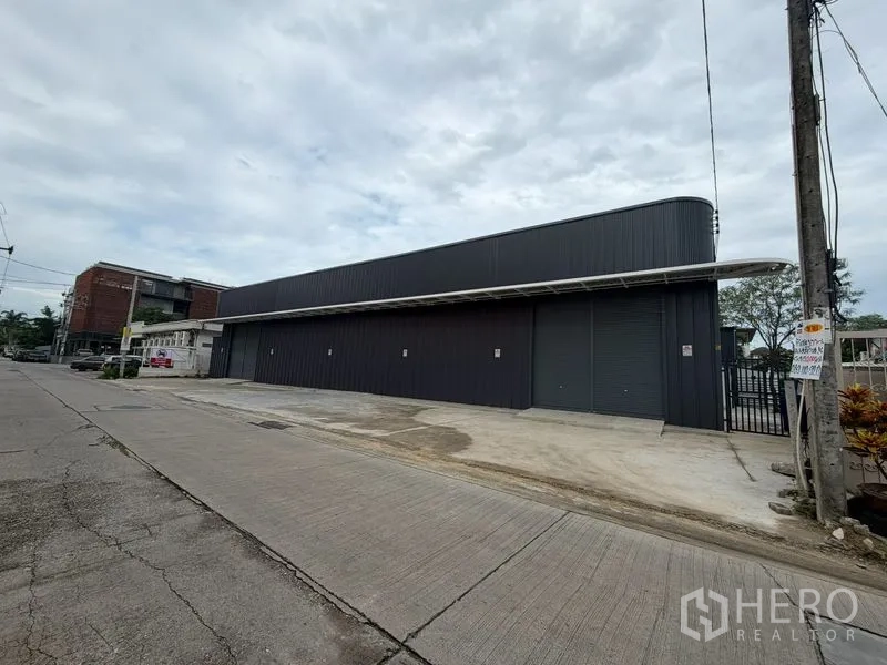 Warehouse for rent in Chatuchak, Bangkok - Street view of a modern warehouse with multiple roll-up doors, a covered loading area, and a gated side entrance.