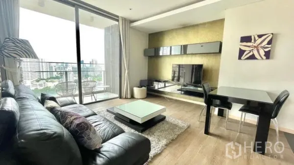Condominium for rent or sale in Watthana, Bangkok - Living room with leather sofa, TV wall, dining table and balcony access.
