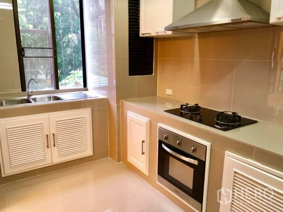 Townhouse for rent in Watthana, Bangkok - Modern closed kitchen with gas hob, built-in oven, double sink and a window with greenery views.