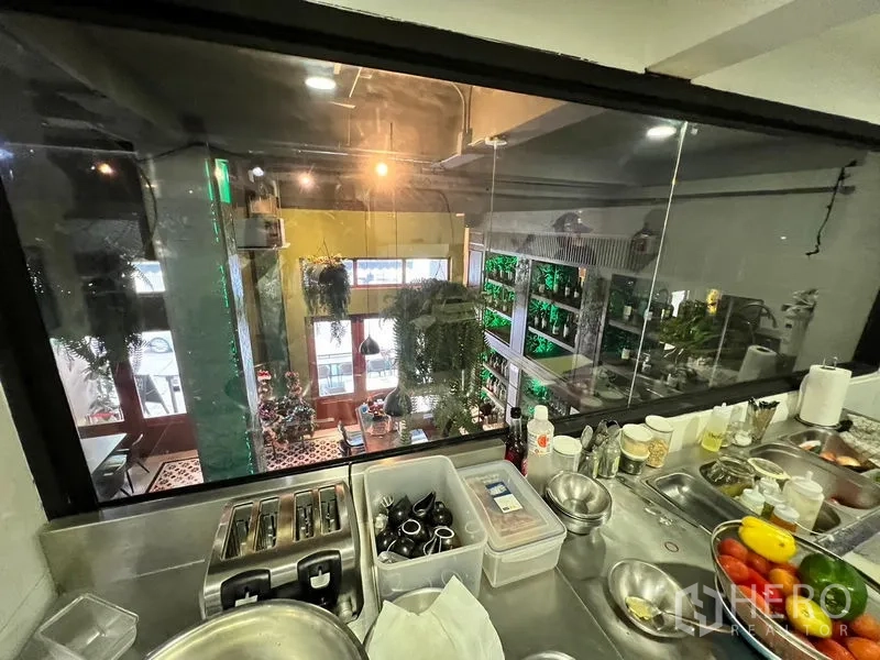 อาคารพาณิชย์ ให้เช่าในWatthana, Bangkok - มุมมองจากห้องเตรียมอาหารผ่านกระจกไปยังโถงรับประทานอาหารที่มีต้นไม้แขวนและประตูไม้สูง.