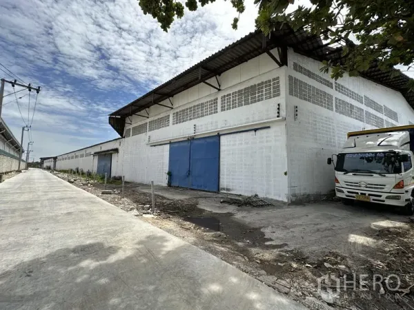 Warehouse for rent in Samut Prakan - Exterior view of a large white warehouse with blue sliding doors, metal roof and a wide concrete access road for trucks.