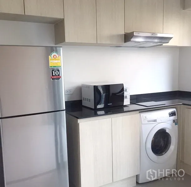 Townhouse for rent in Suan Luang, Bangkok - Contemporary kitchen with light wood cabinets, black countertop, fridge, microwave, washing machine and range hood.
