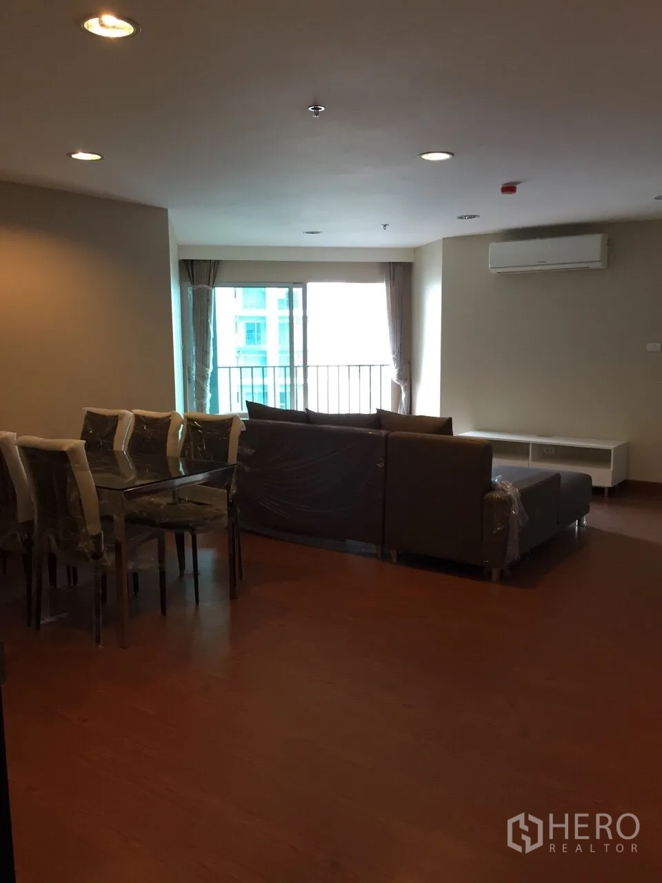 Condominium for rent in Huai Khwang, Bangkok - Open-plan living and dining area with balcony door, air conditioner and wood floors overlooking Bangkok.
