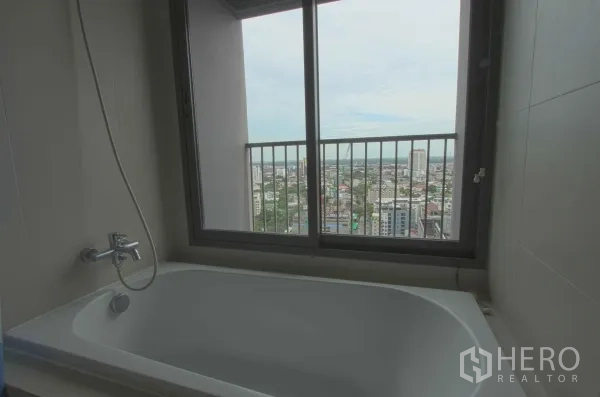 Condominium for rent in Khlong Toei, Bangkok - Bathroom with a soaking tub positioned by a large window offering an open city skyline view.