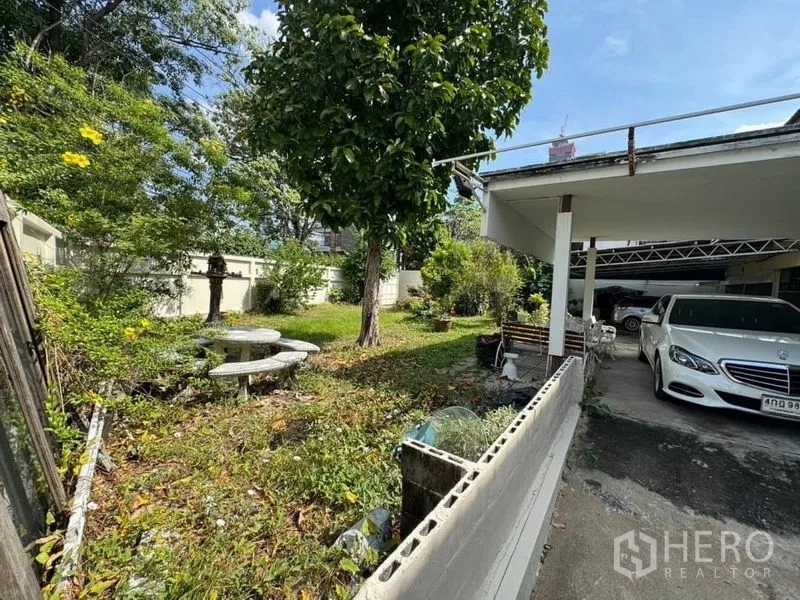 独立式住宅 出租在Watthana, Bangkok - 花园树荫浓密，设有圆形水磨石桌椅，车棚在一侧。