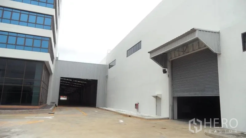 货栈 出租在Bang Sao Thong, Samut Prakan - 仓库外立面，带雨棚的装卸月台、卷闸门与车道，旁有办公楼。