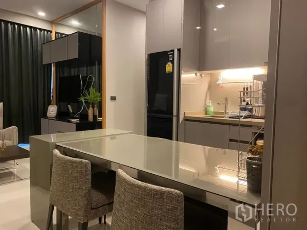 Condominium for rent in Bang Rak, Bangkok - Open-plan kitchen with gray cabinets, island dining table, and a view into the cozy living area.
