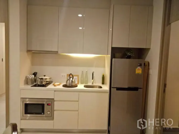 Condominium for rent or sale in Pathum Wan, Bangkok - Modern kitchenette with glossy cabinets, electric cooktop, microwave, sink, and stainless-steel refrigerator.