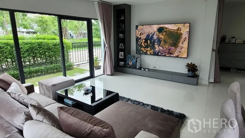 Detached House for rent in Don Mueang, Bangkok - Cozy lounge with sectional sofa, wall-mounted TV and full-height windows to the garden.