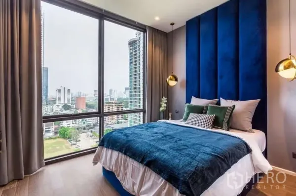 Condominium for rent in Pathum Wan, Bangkok - Corner bedroom with deep blue headboard and floor-to-ceiling windows overlooking the Bangkok skyline.