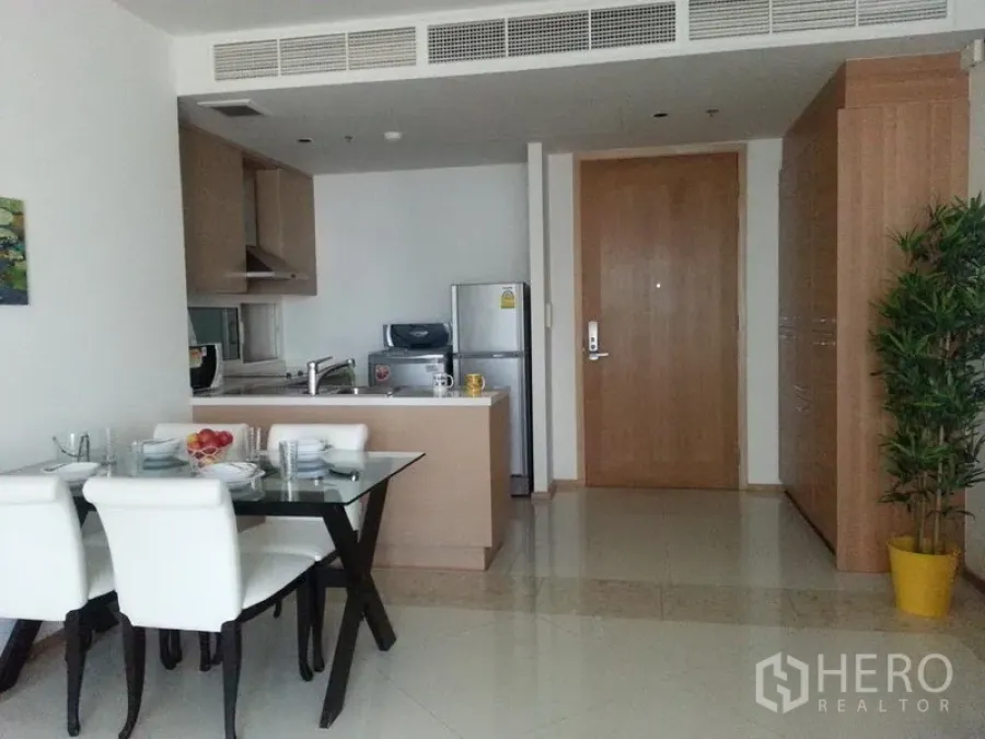 Condominium for rent in Sathon, Bangkok - Open-plan kitchen with light wood cabinets, island, and a glass dining table by the entry in a Sathon condo.