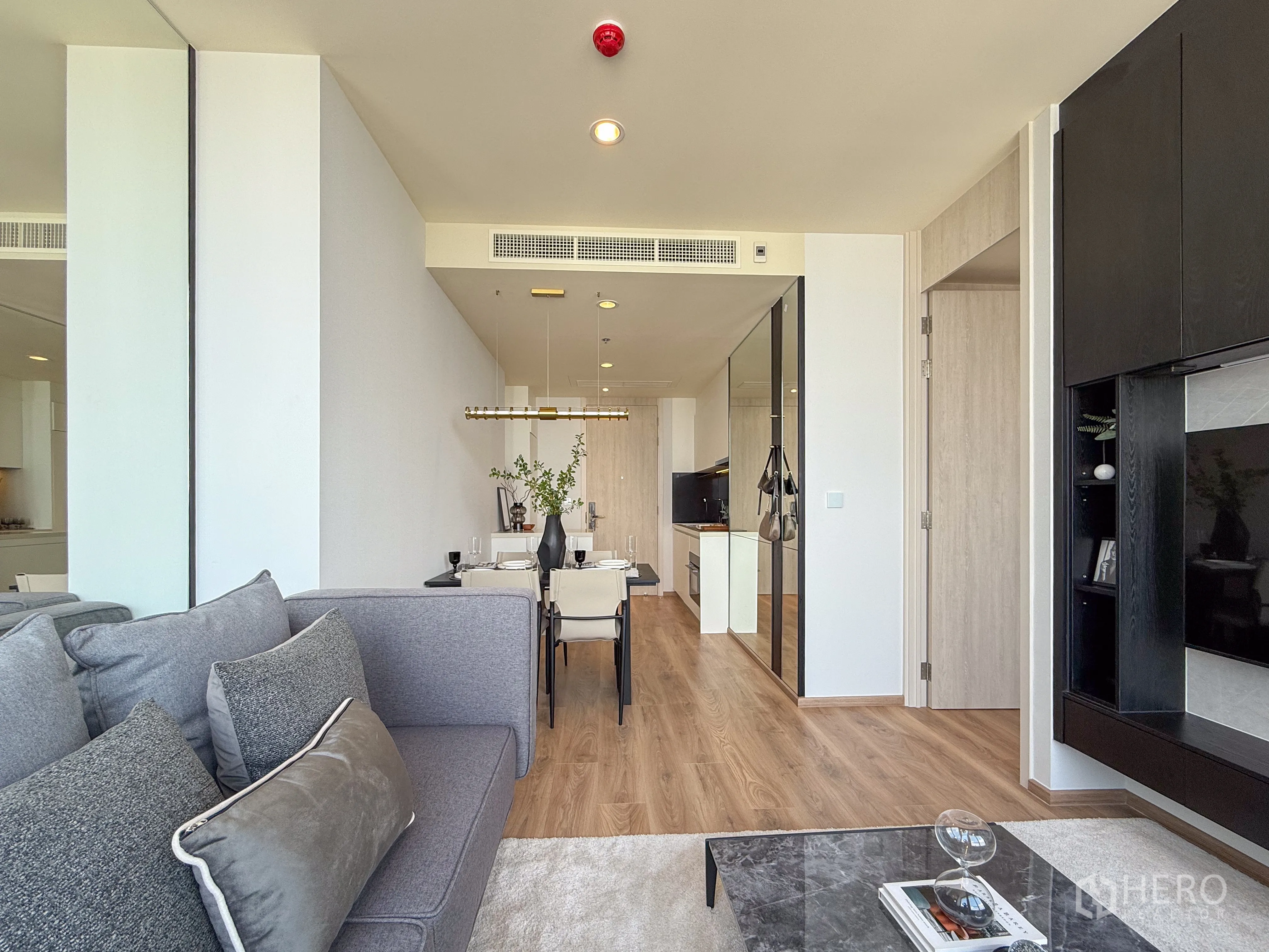 公寓 for sale in Watthana, Bangkok - 现代客厅设灰色沙发、电视墙柜、四人餐桌、木地板与镜面墙，通向厨房。