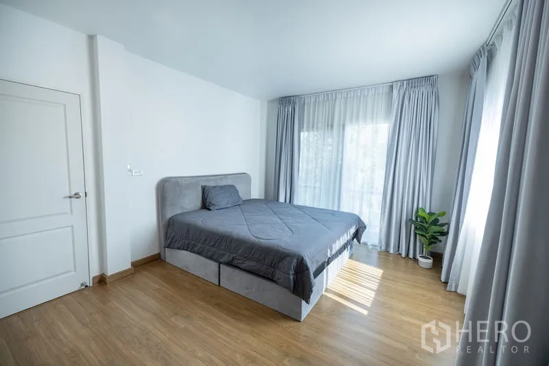 Detached House for rent or sale in Bang Phli, Samut Prakan - Spacious master bedroom with gray bed, corner windows and wood floors.