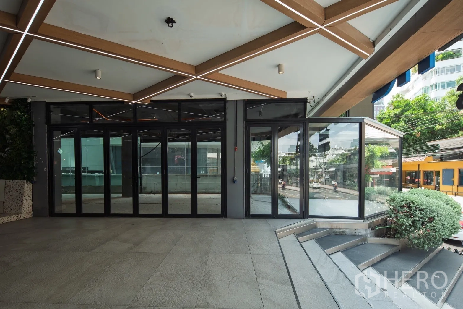 พื้นที่ขายปลีก ให้เช่า - ด้านหน้าร้านชั้นถนน ประตูกระจกเต็มบาน ใต้กันสาดลวดลายเรขาคณิต พร้อมบันไดและพุ่มไม้จัดสวน