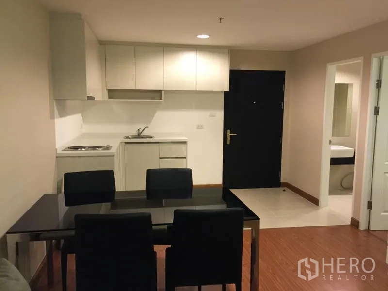 Condominium for rent in Huai Khwang, Bangkok - Compact white kitchenette with cabinets, sink and a dining table by the entry.
