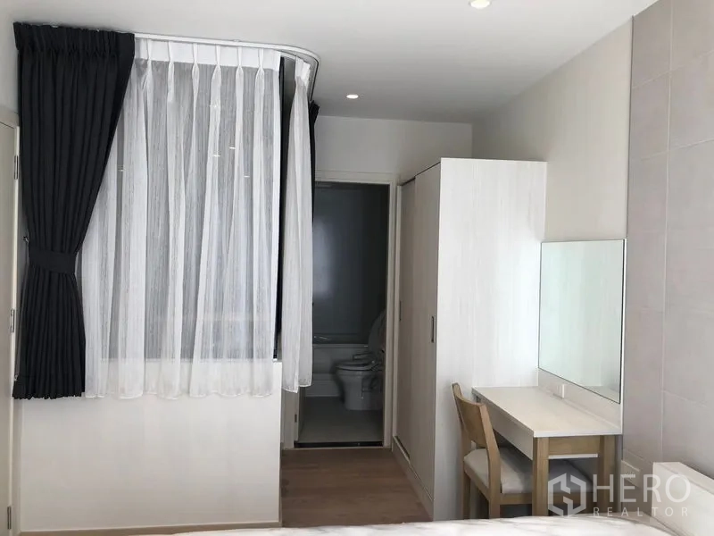 Condominium for rent in Watthana, Bangkok - Bedroom view showing wardrobe, vanity, sheer and blackout curtains and bathroom doorway.