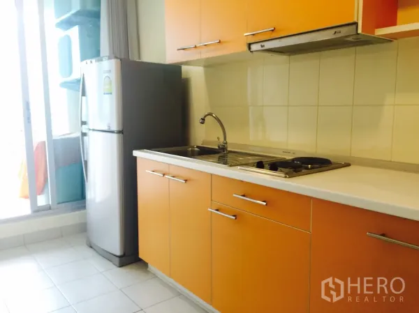 Condominium for rent in Watthana, Bangkok - Separate kitchen with orange cabinets, sink, electric cooktop, and refrigerator beside the balcony door.