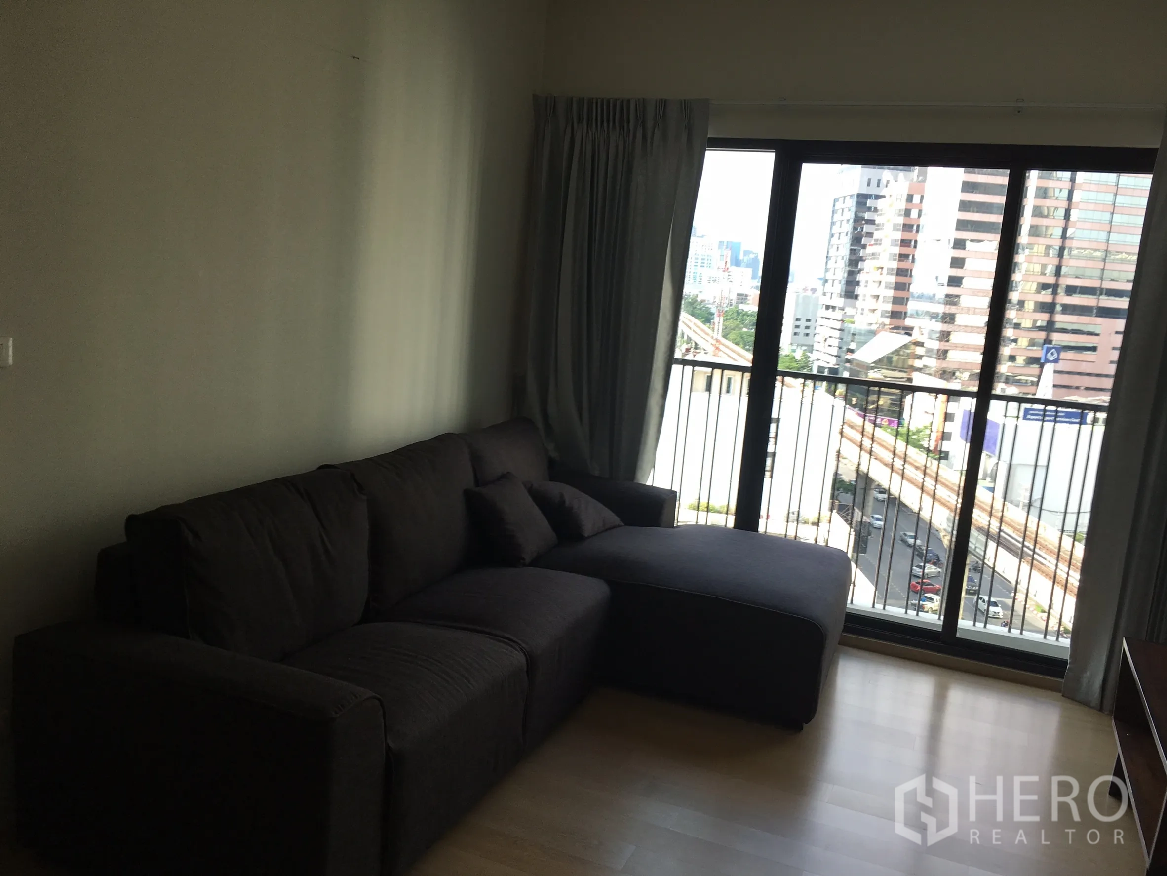 คอนโด ให้เช่าในPhaya Thai, Bangkok - ห้องนั่งเล่นมีโซฟาเข้ามุมสีเข้ม เปิดสู่ระเบียงชมวิวเมือง ใกล้รถไฟฟ้า