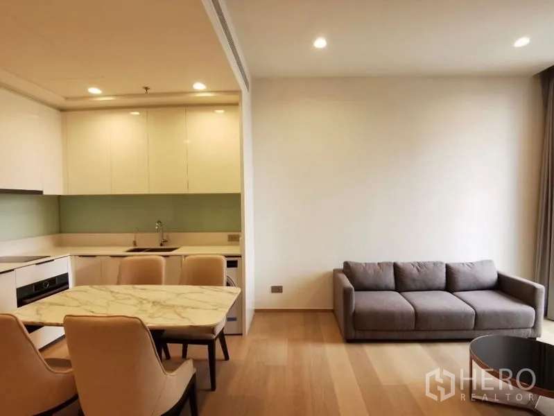 公寓 出租在Bang Rak, Bangkok - 开放式客厅与L形厨房，灰色沙发与石面餐桌，木地板灯光明亮。