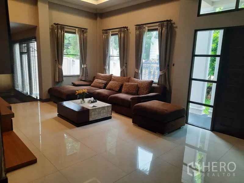 Detached House for rent in Khlong Toei, Bangkok - Bright living room with an L-shaped sofa, large windows and glossy tiled floor.
