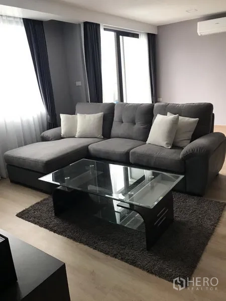 公寓 出租在Bang Rak, Bangkok - 采光良好的客厅，灰色L形沙发、玻璃茶几、地毯与大窗帘窗。