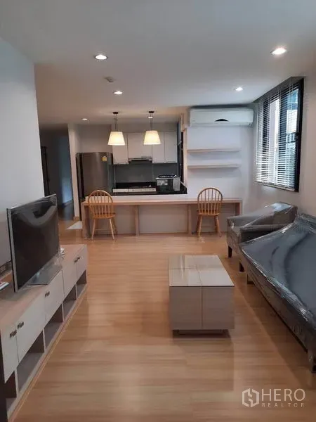 Condominium for rent in Watthana, Bangkok - Bright living room with TV console, sofa and coffee table on wood floors, opening to a kitchen with breakfast bar and pendant lights.