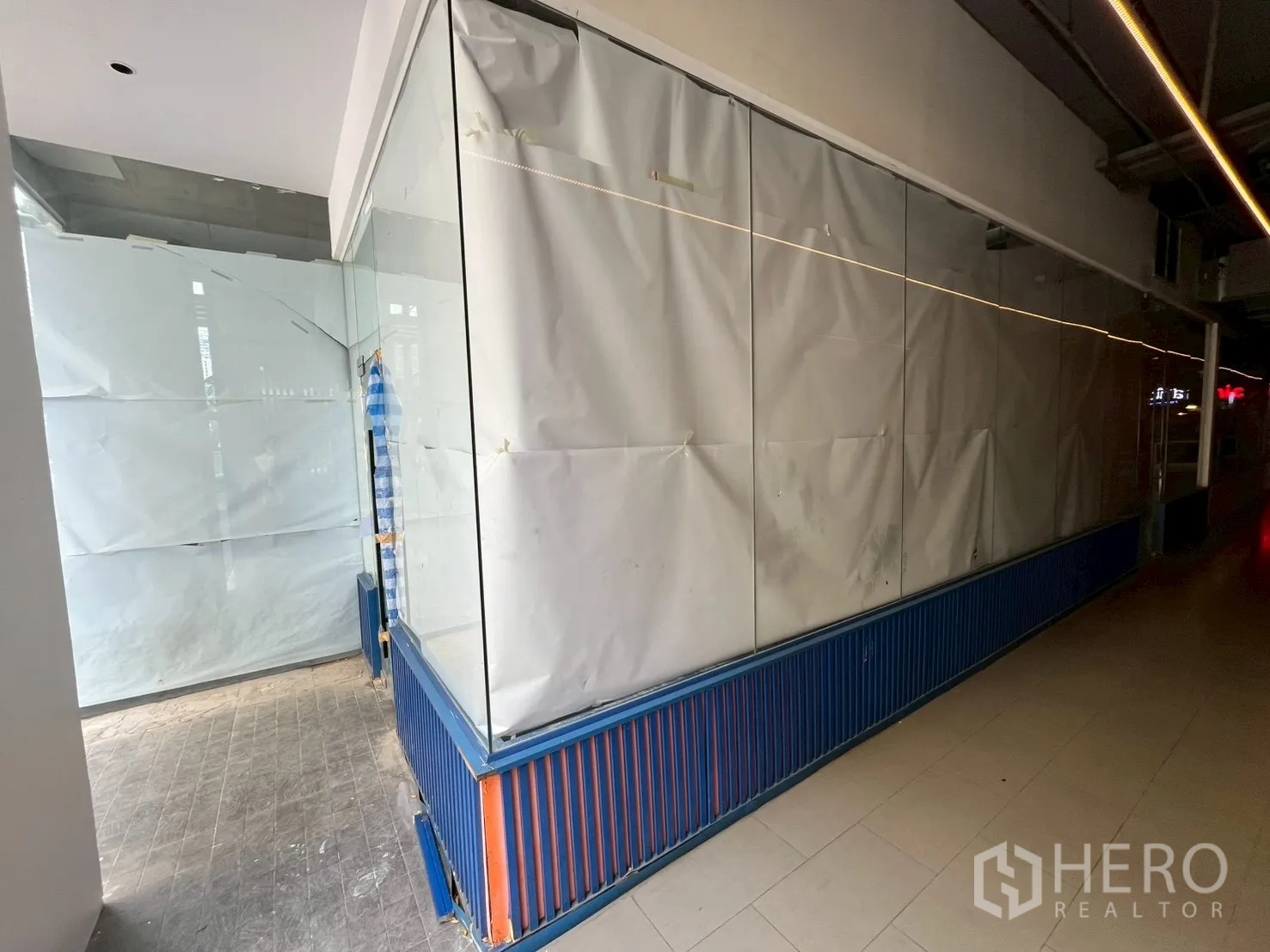 Retail Space for rent in Watthana, Bangkok - Long glass-front retail unit with paper‑covered windows, blue slatted kickboard, tiled floor, and linear ceiling lights along an indoor corridor.