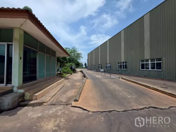 Factory for rent in Mueang Samut Sakhon, Samut Sakhon - Entrance road between a glass‑front office and large factory halls with ramp access under a clear sky.