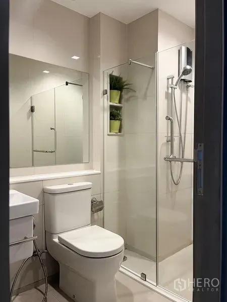 Condominium for sale in Ratchathewi, Bangkok - Modern bathroom featuring a glass-enclosed shower, toilet, and wide mirror above a vanity.