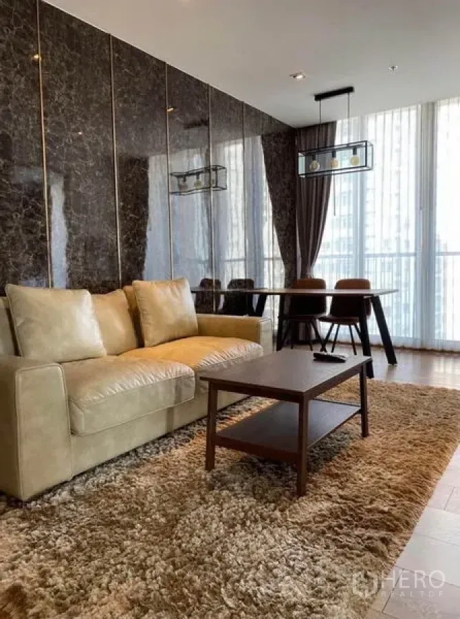 Condominium for rent in Khlong Toei, Bangkok - Elegant living room with marble feature wall, beige sofa, shag rug, and dining set by floor-to-ceiling windows.