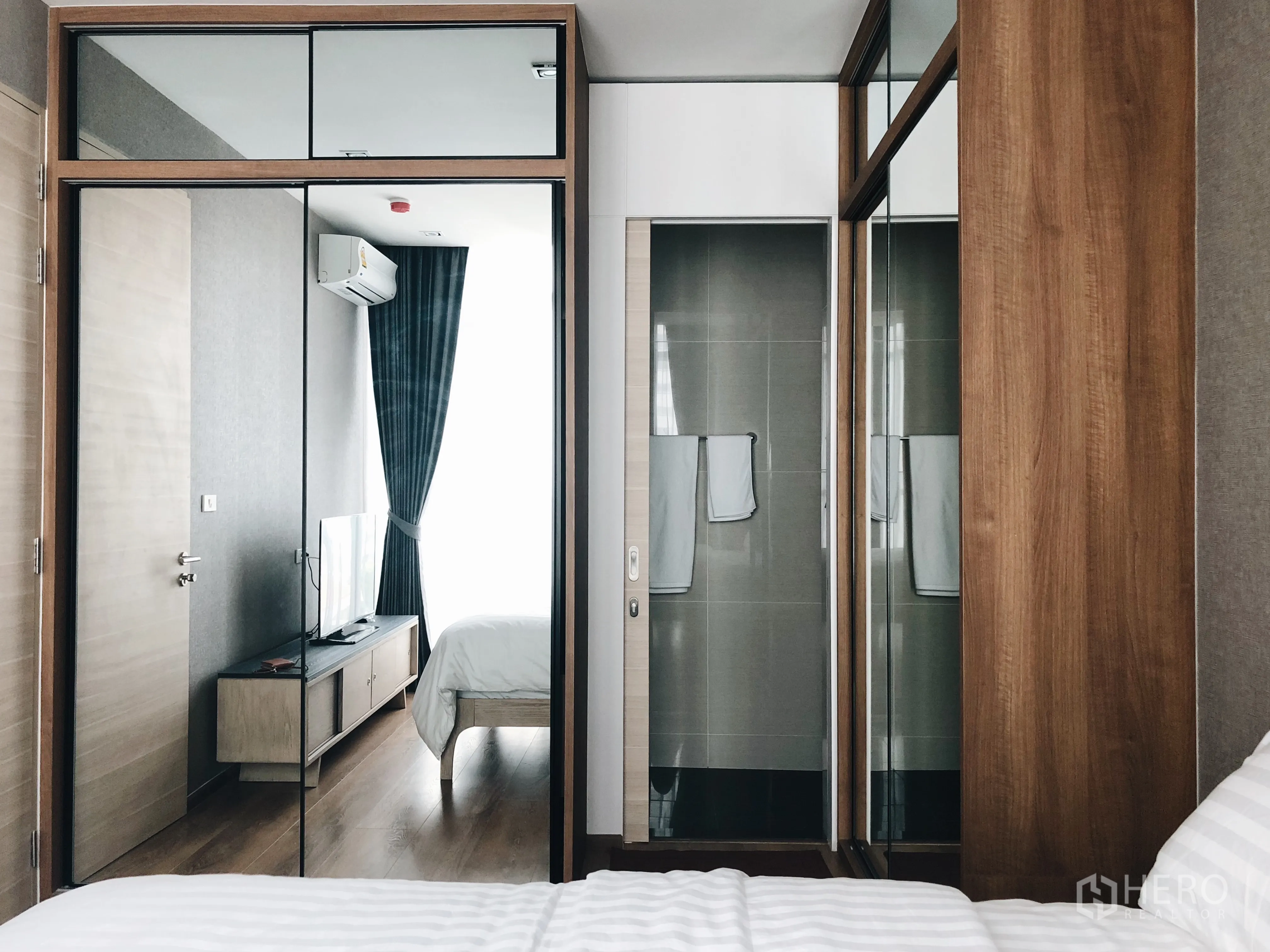 Condominium for rent in Khlong Toei, Bangkok - Bedroom with mirrored built-in wardrobe and sliding door to tiled bathroom, warm wood accents.