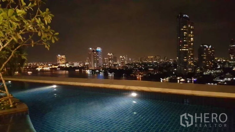 Condominium for rent in Bang Kho Laem, Bangkok - Nighttime pool with illuminated water and city skyline across the river.
