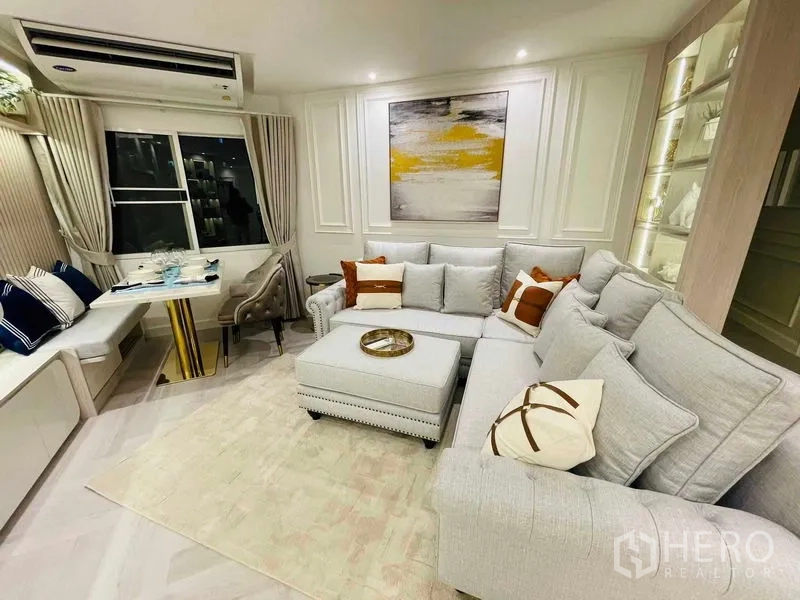 Condominium for sale in Yan Nawa, Bangkok - Bright living room with L-shaped gray sofa, ottoman, wall art, and window seating.