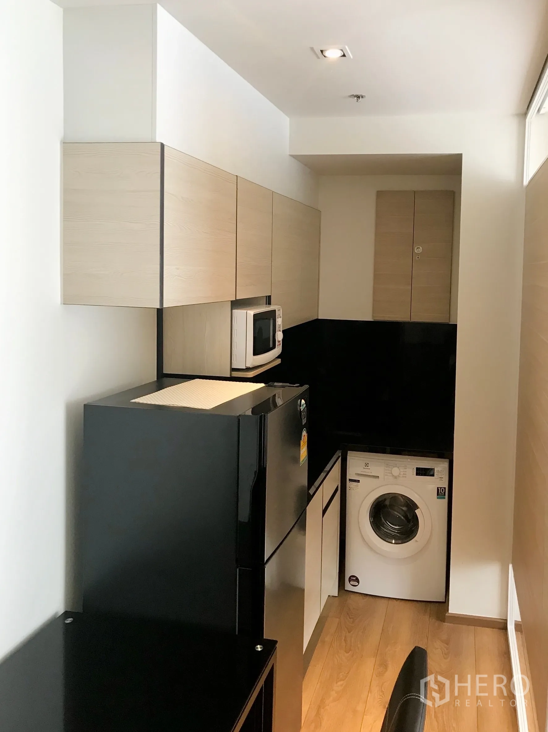 Condominium for sale in Khlong Toei, Bangkok - Compact kitchenette with light wood cabinets, a black refrigerator, microwave, and washing machine.
