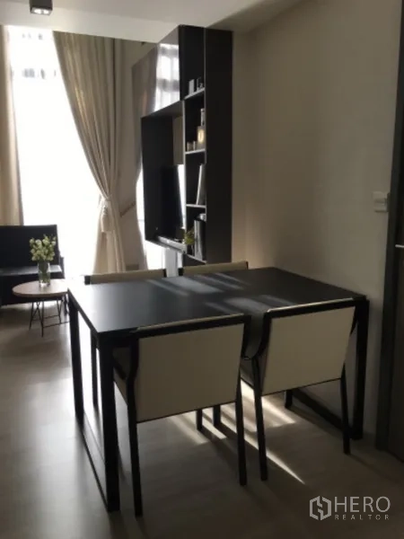 Condominium for sale in Pathum Wan, Bangkok - Bright dining area with a black table and two chairs beside an open-shelf media wall and sheer curtains.