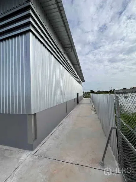 Warehouse for rent in Phra Samut Chedi, Samut Prakan - Side walkway along a corrugated metal-clad warehouse with chain-link fence and overhanging roof.