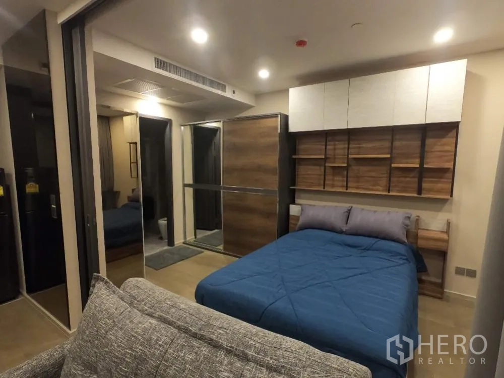Condominium for rent in Watthana, Bangkok - Cozy bedroom with blue duvet, gray sofa at the foot, mirrored closet doors, and doorway to the bathroom.