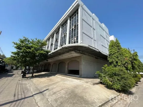 店屋 出租在Khlong Toei, Bangkok - 多层商铺建筑外观，宽大雨棚、数个卷帘门车间，带坡道与街边绿化。