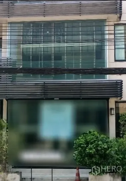 零售空间 出租 - 位于曼谷Wang Thonglang的三层现代临街店面，整面落地玻璃立面与黑色百叶遮阳，前方有绿化与电线。