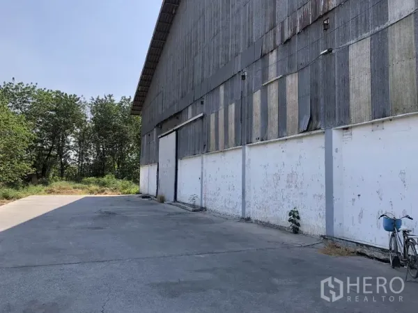 Warehouse for rent in Nonthaburi - Side view of the warehouse facade with a long concrete apron and greenery.