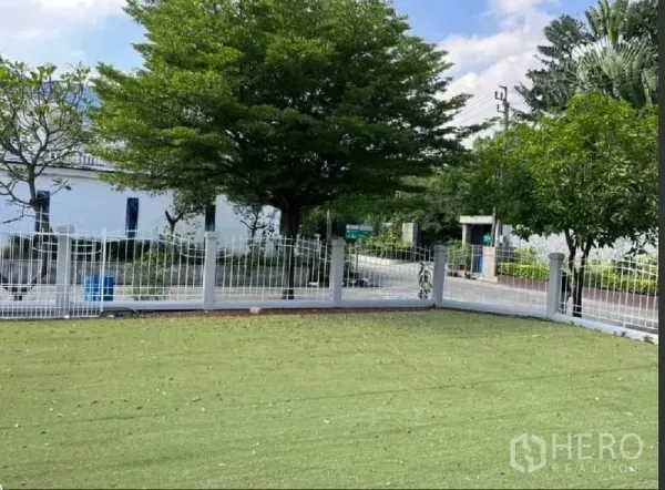 独立式住宅 出租在Samut Prakan - 转角草坪与白色金属栅栏，临静谧绿荫街道。