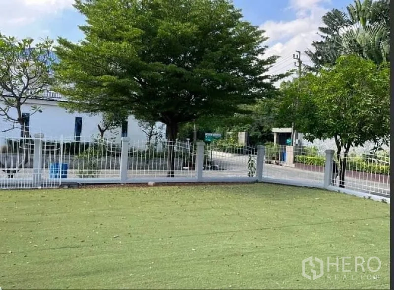 独立式住宅 出租在Samut Prakan - 转角草坪与白色金属栅栏，临静谧绿荫街道。