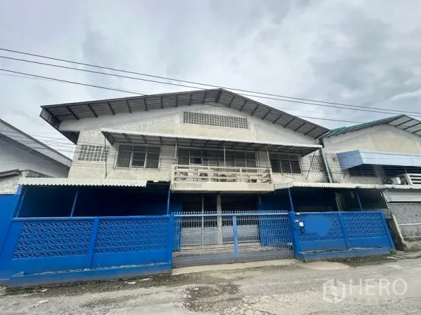Factory for rent in Lam Luk Ka, Pathum Thani - Front elevation of the factory with blue fence and sliding gate.