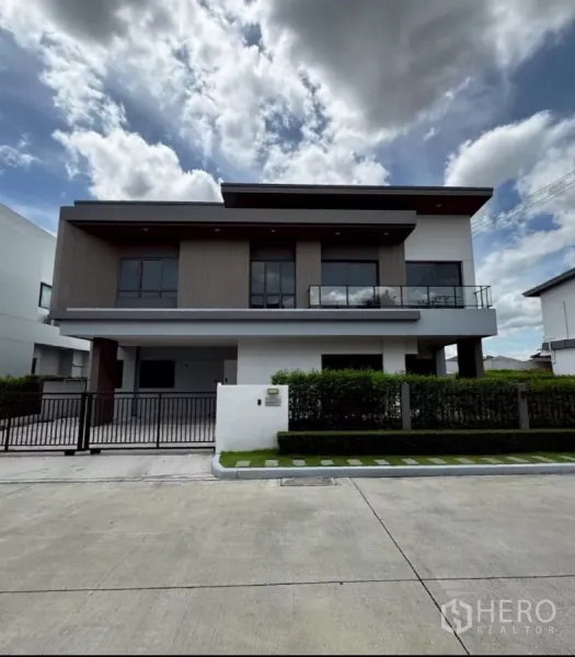 บ้าน ให้เช่าหรือขายในLat Krabang, Bangkok - บ้านเดี่ยวสไตล์โมเดิร์น 2 ชั้น ระเบียงกระจก ประตูรั้วหน้าบ้าน และแนวพุ่มไม้ตัดแต่งเรียบร้อยใต้ท้องฟ้ามีเมฆ.