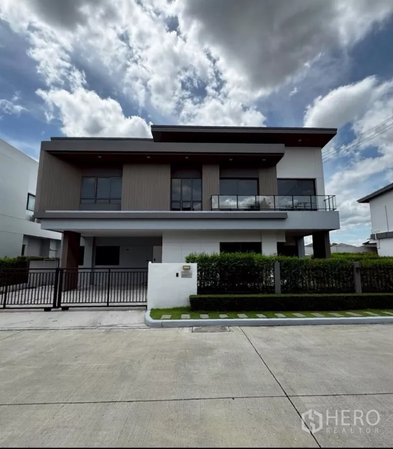 บ้าน ให้เช่าหรือขายในLat Krabang, Bangkok - บ้านเดี่ยวสไตล์โมเดิร์น 2 ชั้น ระเบียงกระจก ประตูรั้วหน้าบ้าน และแนวพุ่มไม้ตัดแต่งเรียบร้อยใต้ท้องฟ้ามีเมฆ.