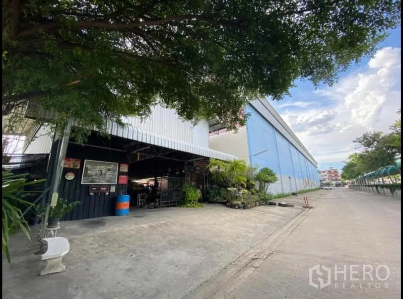 货栈 出租在Sai Mai, Bangkok - 仓库入口设钢结构雨棚，小型办公区与宽阔混凝土场地，被大树遮荫。