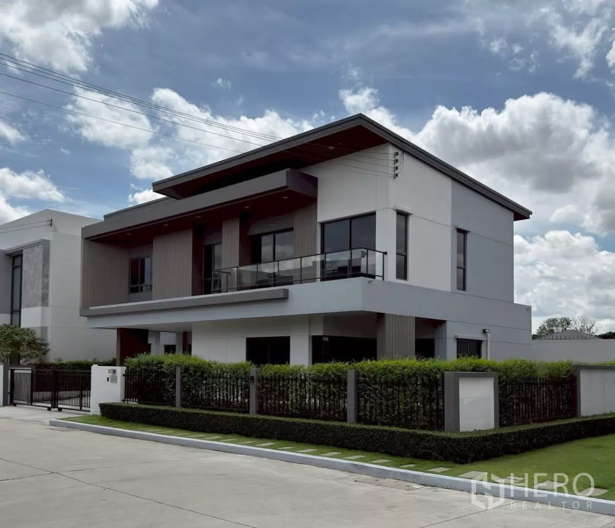 บ้าน ให้เช่าหรือขายในLat Krabang, Bangkok - มุมด้านข้างของบ้านเดี่ยวพร้อมระเบียงราวกระจก กระจกบานใหญ่ และรั้วพุ่มไม้จัดสวน.