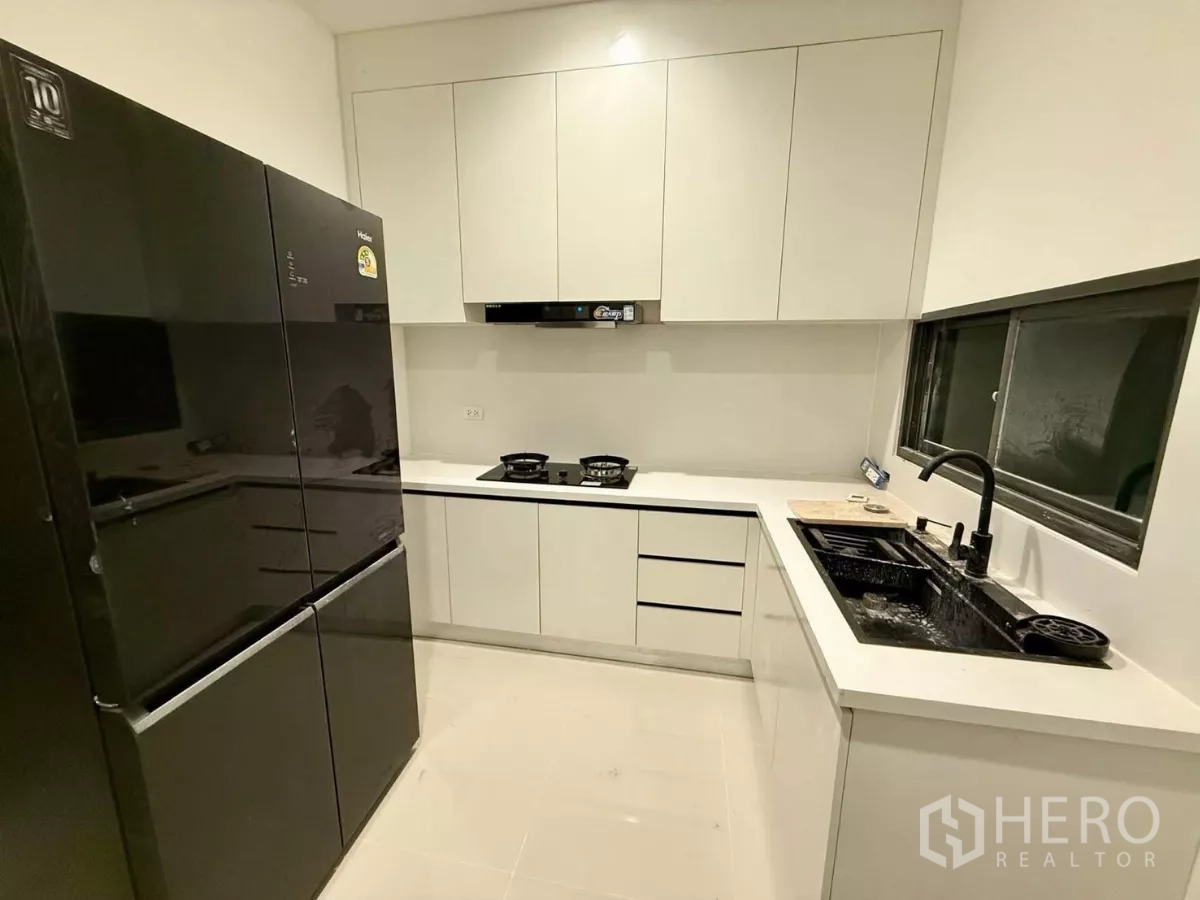 Detached House for rent in Bang Phli, Samut Prakan - Separate L-shaped kitchen with black double-door fridge, gas hob and white cabinets.