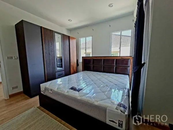 Detached House for rent or sale in Pathum Thani - Corner bedroom featuring a dark wood wardrobe, upholstered headboard and twin windows.